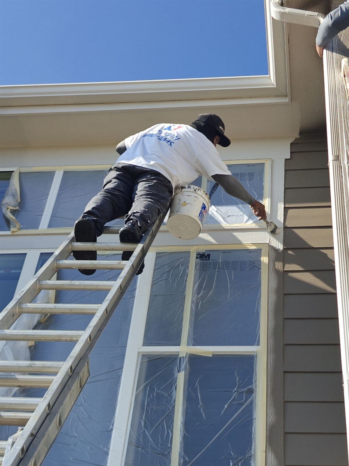 Painter on a ladder applying trim paint along a window and roofline